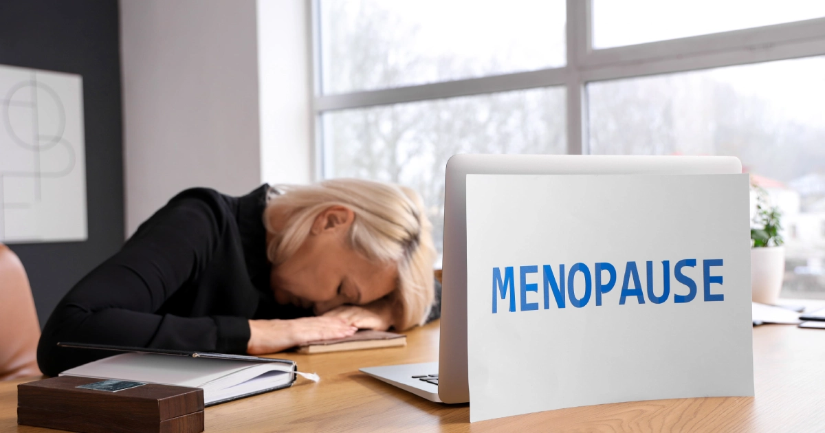 A woman resting her head on a desk beside a laptop labeled ‘MENOPAUSE,’ highlighting fatigue and Somatic-Concentric Sex Therapy for Menopause in Lafayette, CO